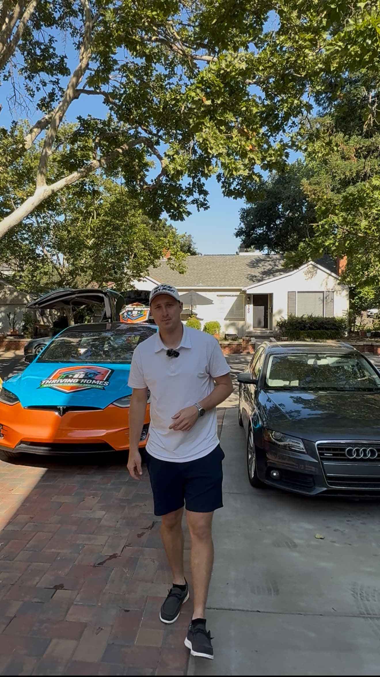 Man in casual attire standing in driveway with a blue and orange car and a black Audi.