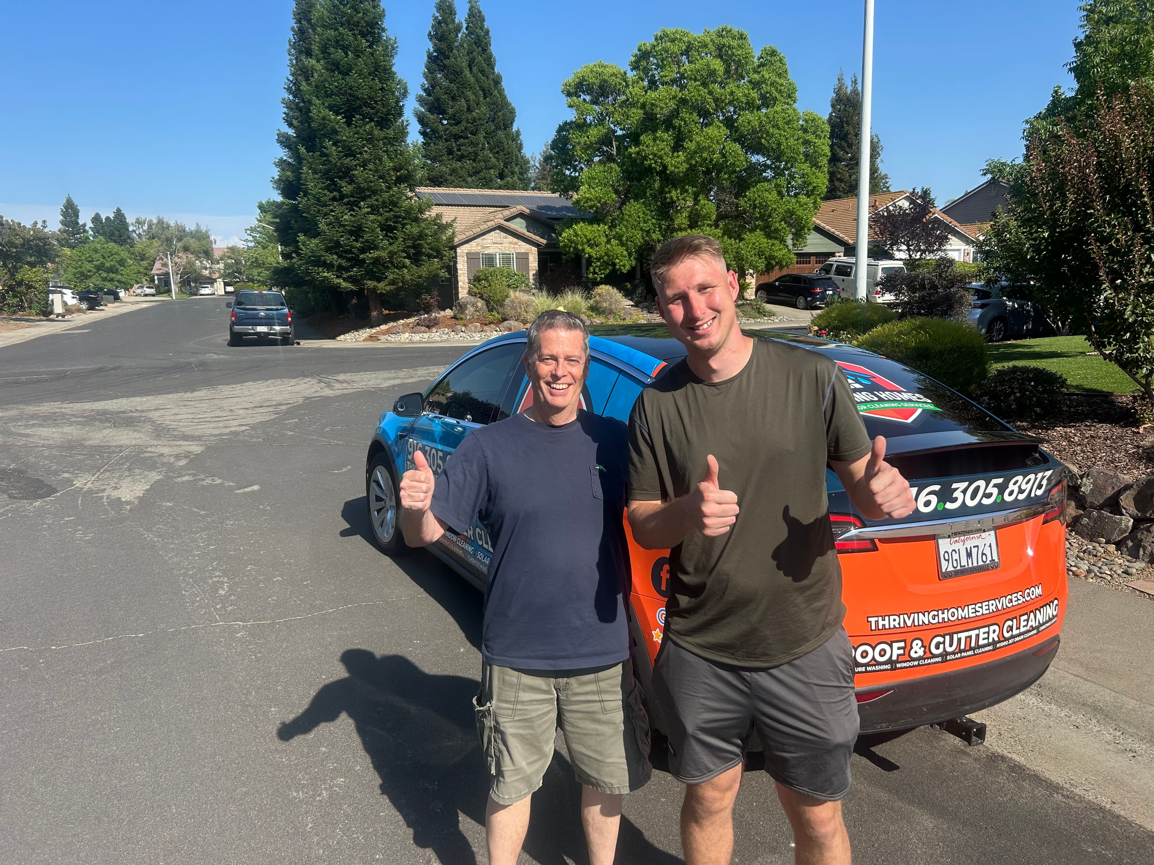 Two men giving thumbs up next to a bright blue and orange service vehicle on a suburban street.