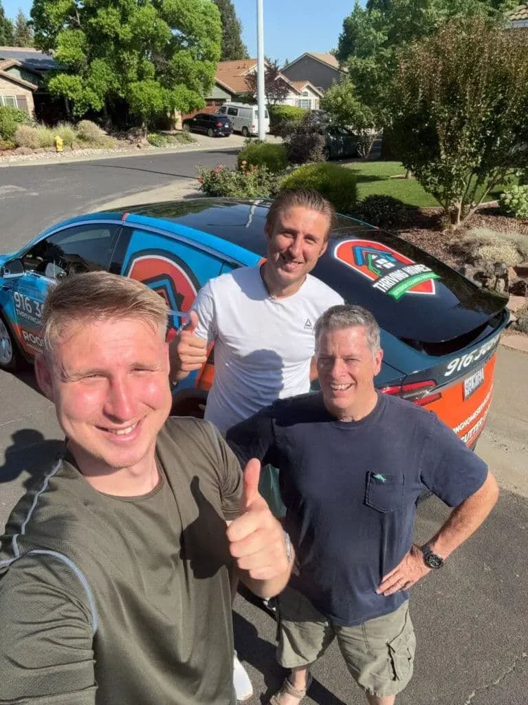 Three men giving thumbs up in front of a colorful car on a sunny neighborhood street.